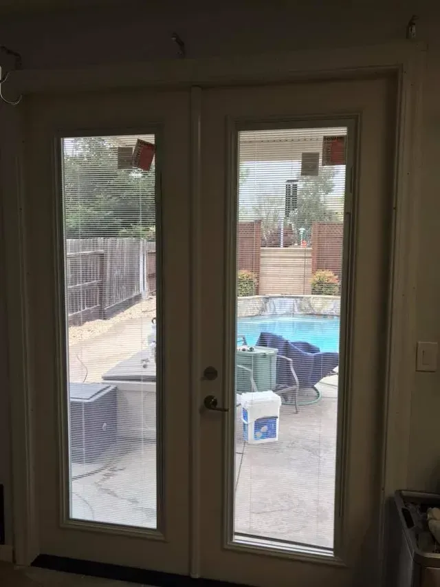 Two glass doors with blinds overlooking a patio with a pool, fence, and furniture.