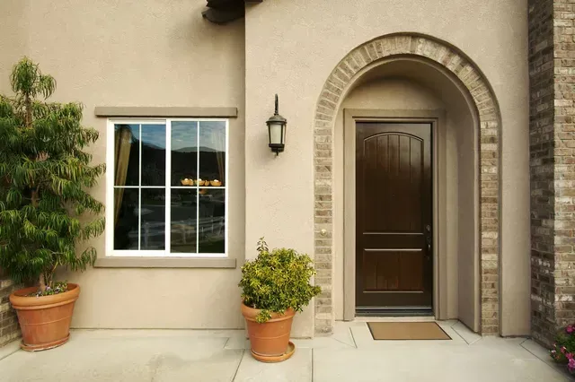 Tan stucco facade with arched doorway and window, flanked by potted plants. Brown door.