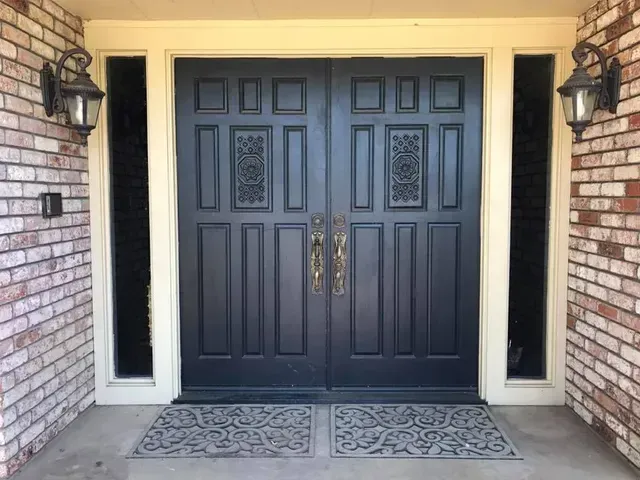 Black double doors with gold handles and decorative carvings, flanked by sidelights and brick.