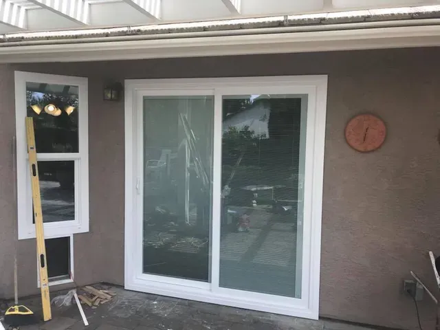 White-framed sliding glass door and adjacent window on brown stucco wall. A level leans against the window.