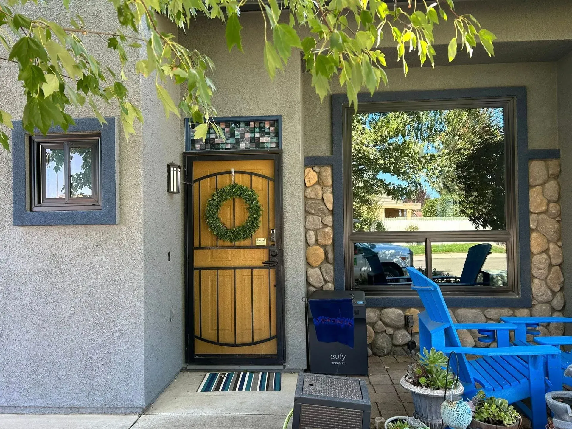 Front exterior of house with light brown door, green wreath, stone accents, blue chairs, and a window reflecting trees.