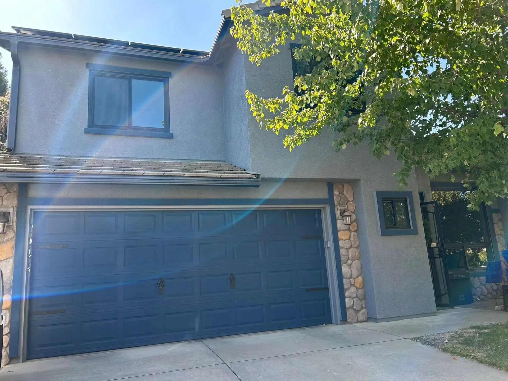 Two-story house with a blue garage door, gray stucco, and stone accents. A tree is to the right.