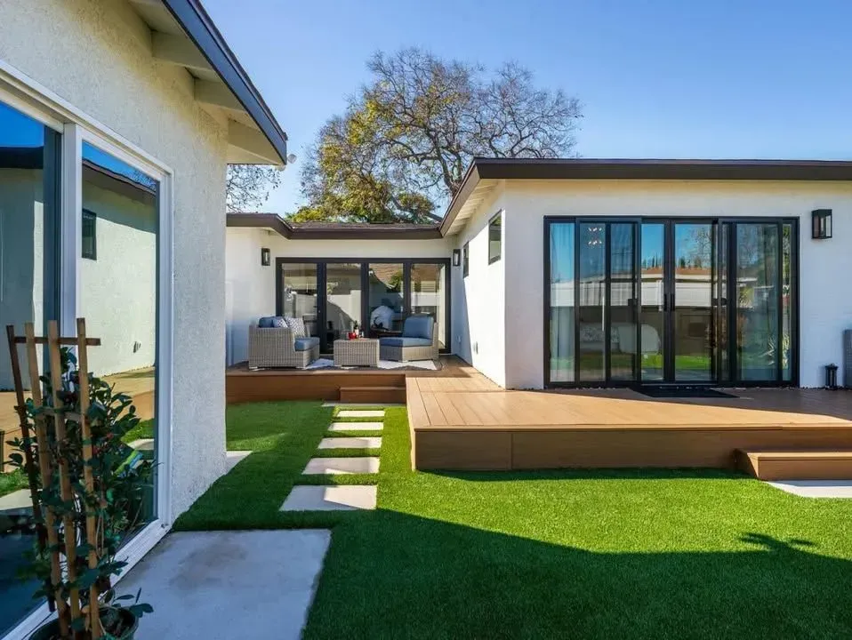 Backyard with artificial grass, wooden deck, stepping stones, and modern white house.