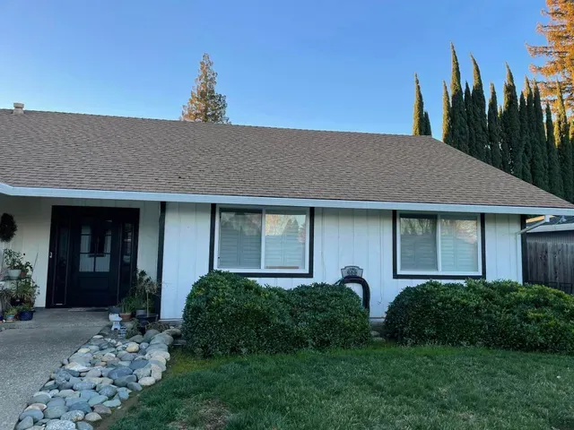 White house with brown roof, black trim around windows and door. Green bushes and grass in front.