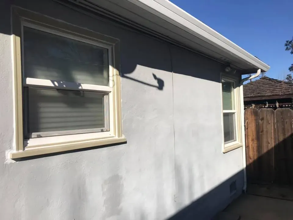 Exterior of a light gray stucco building with two windows. The sky is blue. A wooden fence is on the right.