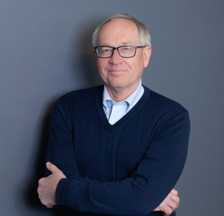Portrait of a man with glasses in a navy sweater, standing with arms crossed against a gray background