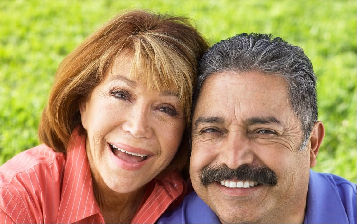 Two smiling people outdoors, close together in front of green grass.