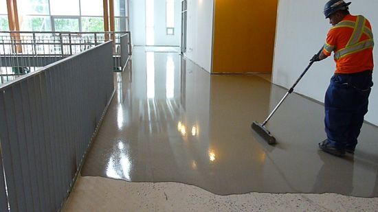 Worker in orange vest spreading wet floor coating in a bright indoor hallway