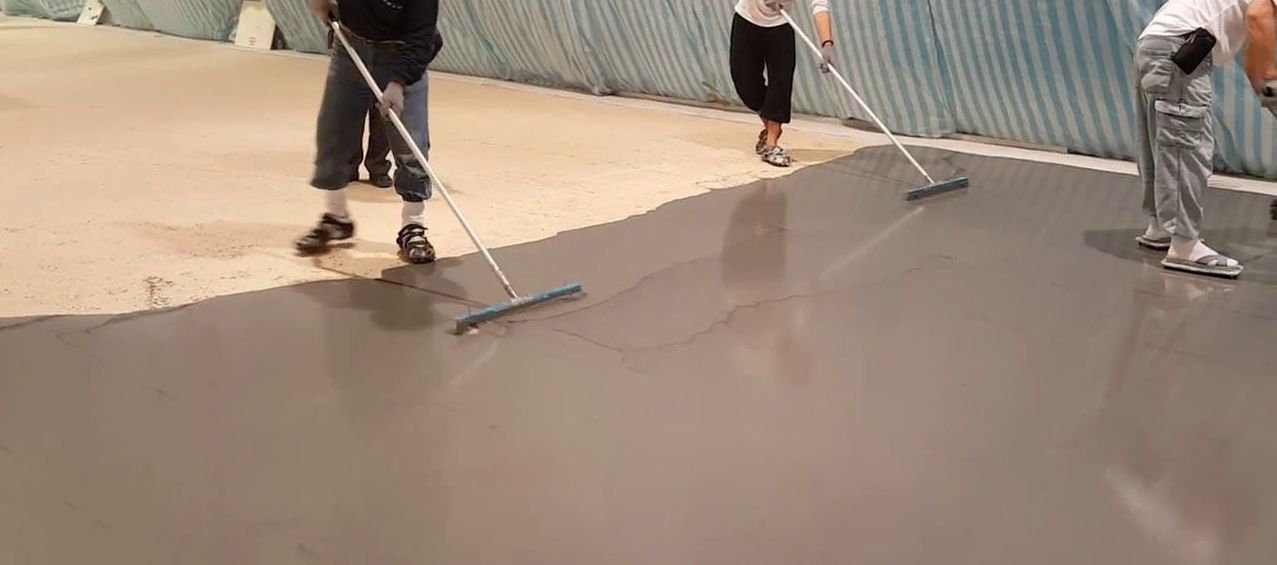 Workers smoothing wet concrete on a driveway, using squeegees beside a corrugated metal wall.