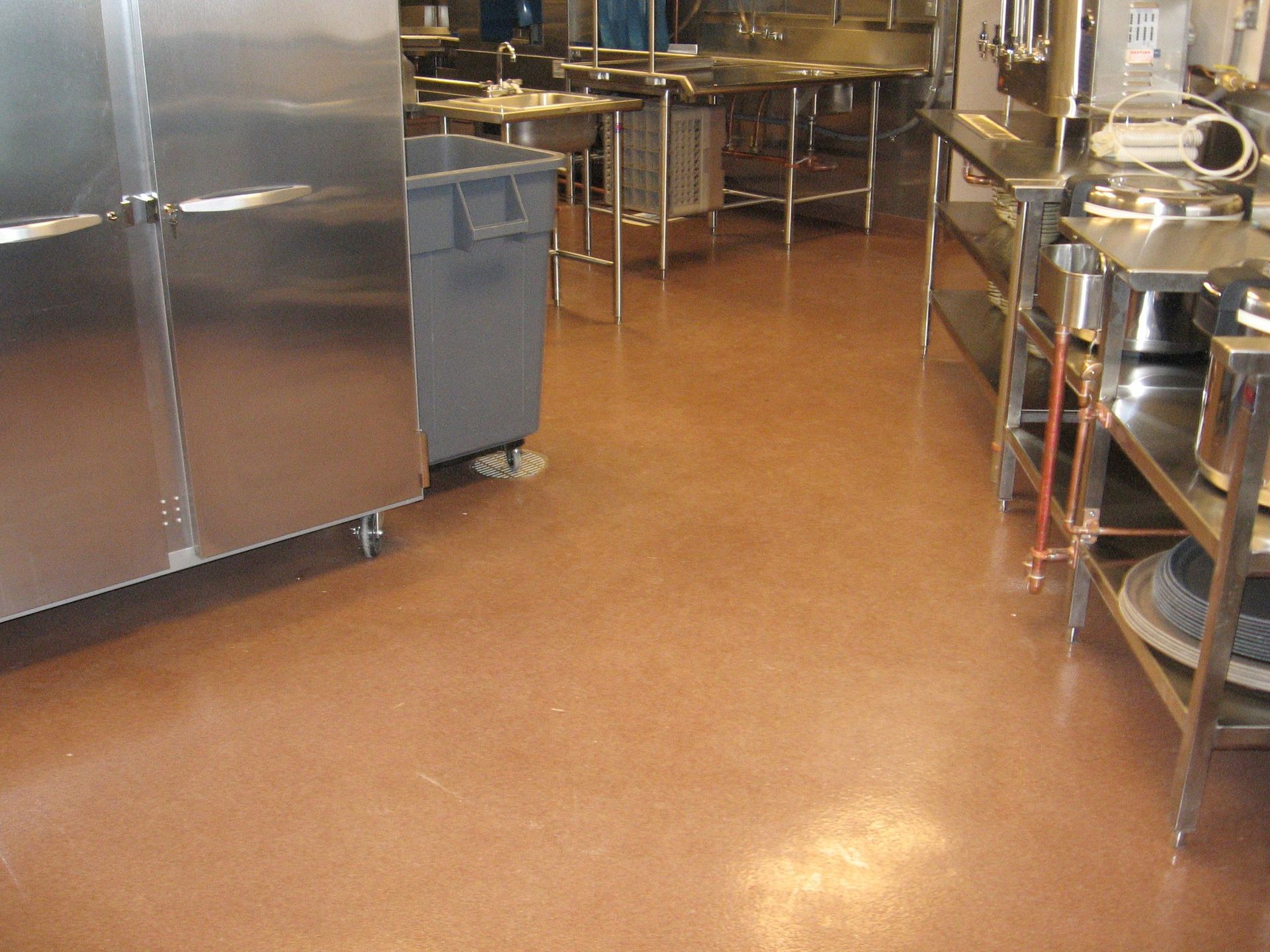 Empty commercial kitchen with stainless steel counters, shelves, and a brown floor