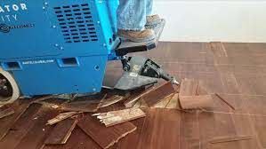 Blue machine on a wooden floor amid scattered broken boards and debris, suggesting repair or cleanup work