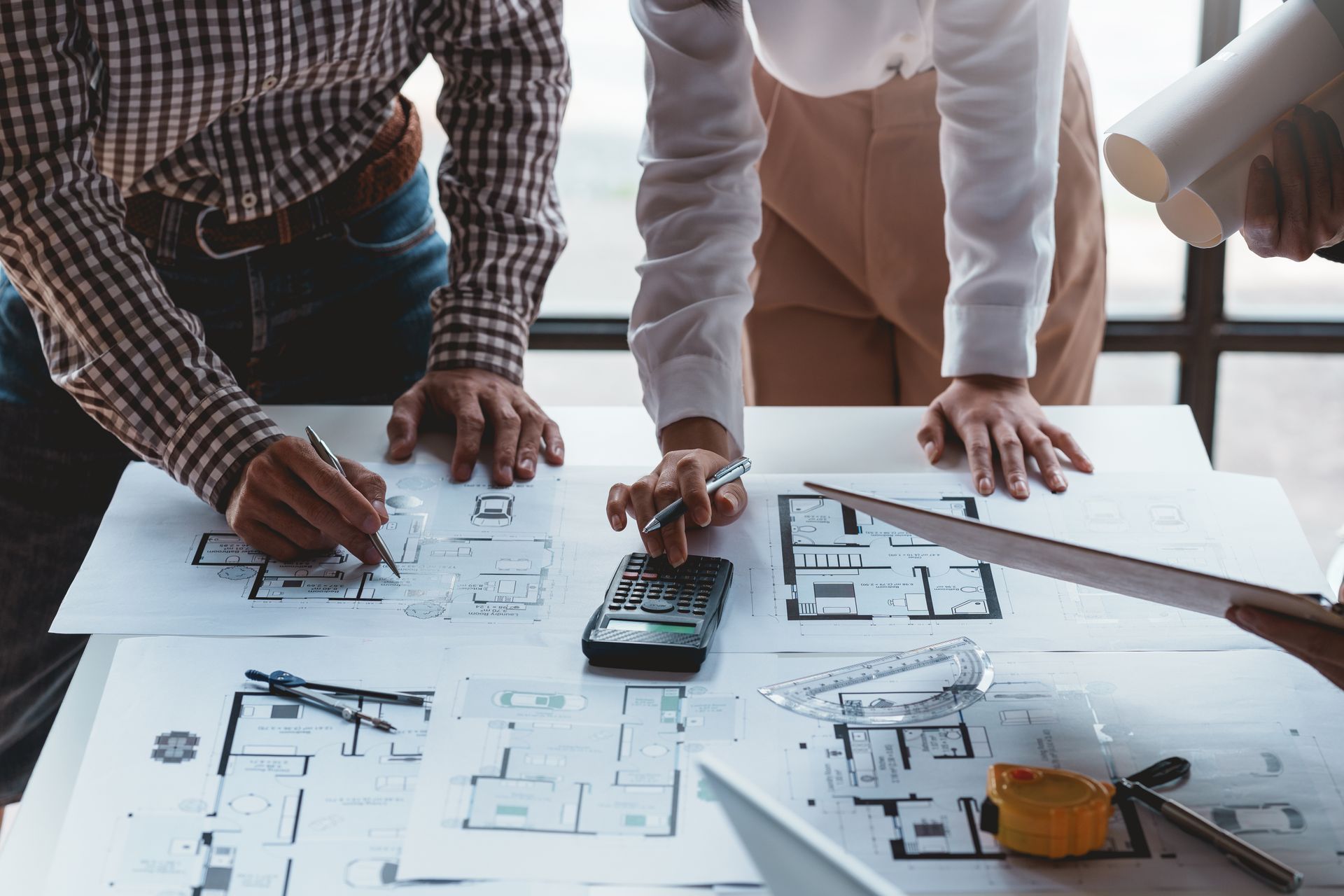 Contractors reviewing home addition blueprints with a calculator and drafting tools Contractors reviewing home addition blueprints with a calculator and drafting tools