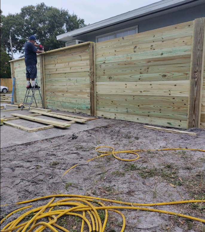 Worker installing a wooden privacy fence beside a house, with lumber, ladder, and coiled hose in the yard.
