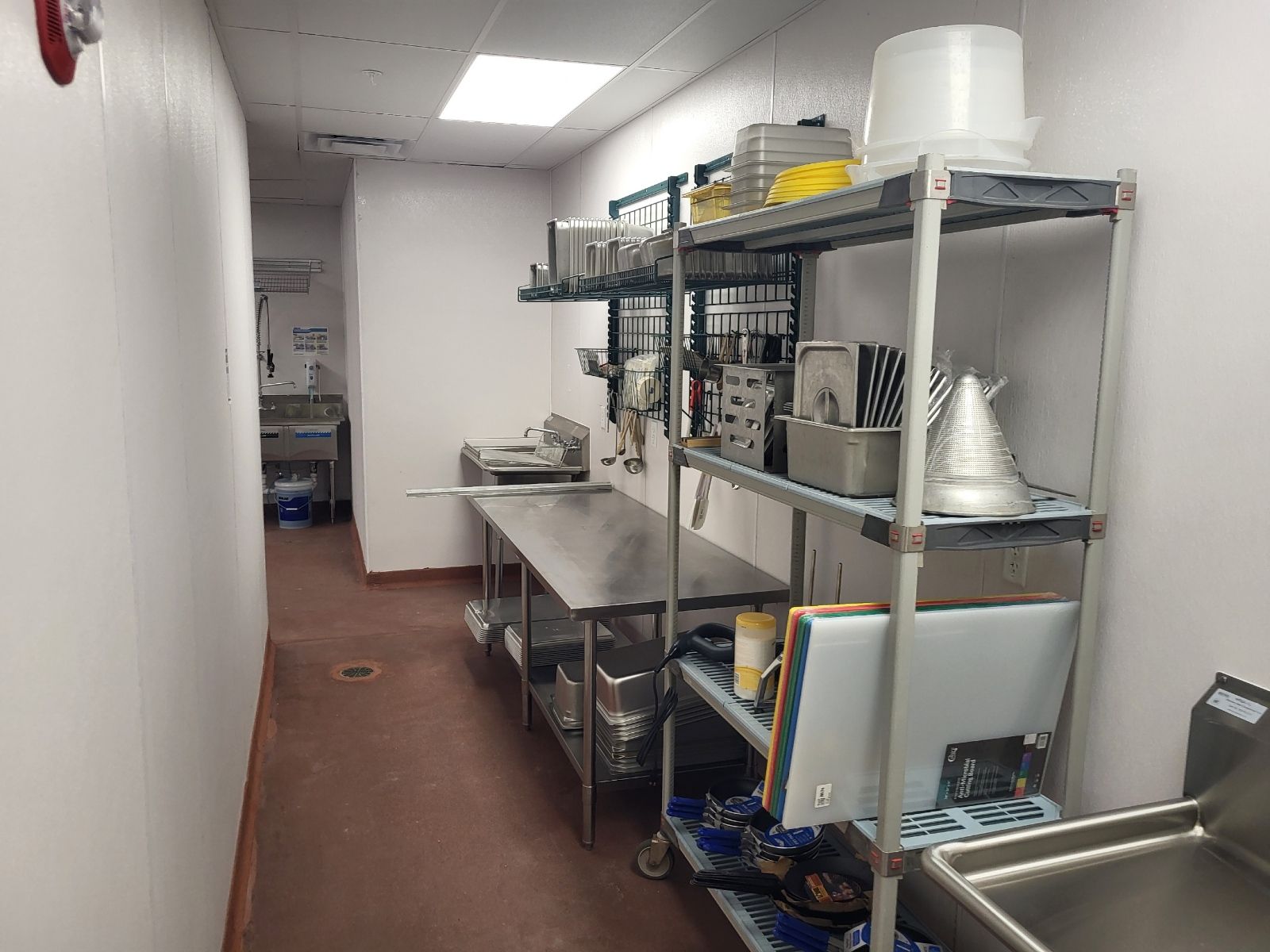 Narrow commercial kitchen corridor with metal prep tables, shelving, buckets, and stainless equipment