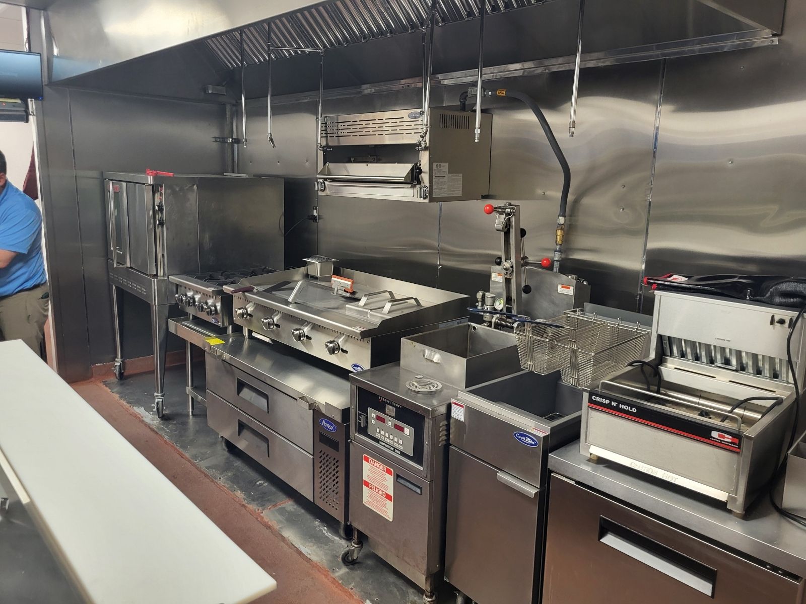 Commercial kitchen with stainless steel counters, ovens, sinks, and cooking equipment under bright hood vents