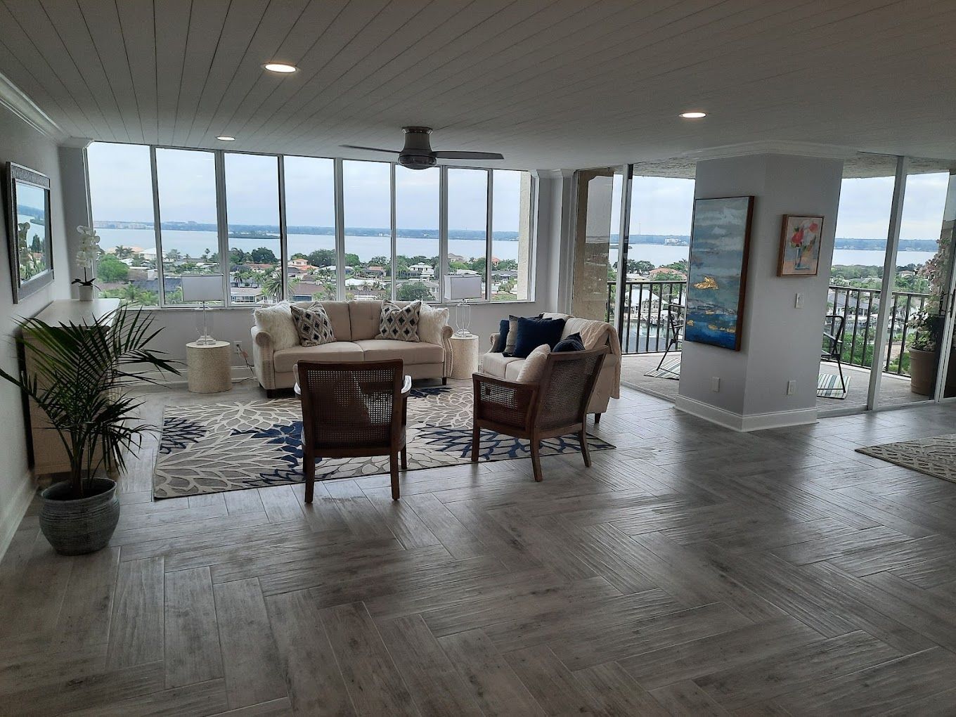Living room with water view, beige couch, rattan chairs, blue rug, and large windows.