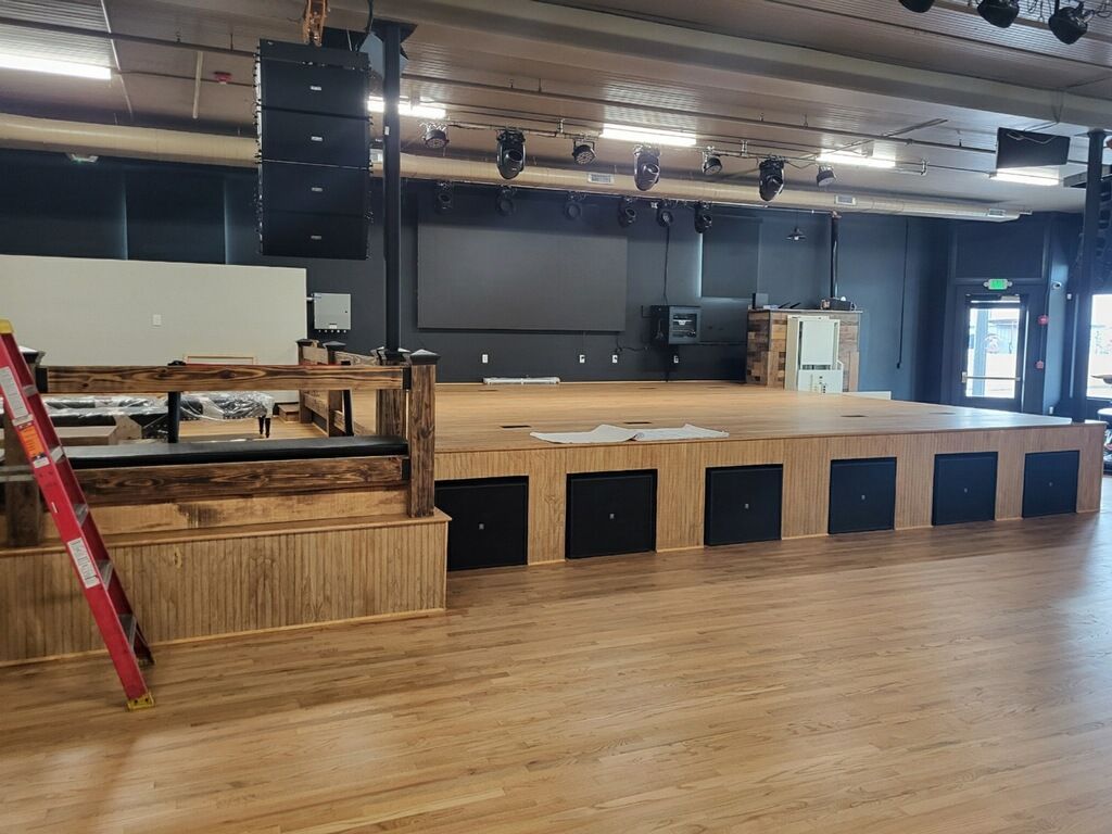 Empty event hall with wooden floor, stage, black speakers, and a red ladder on the left.