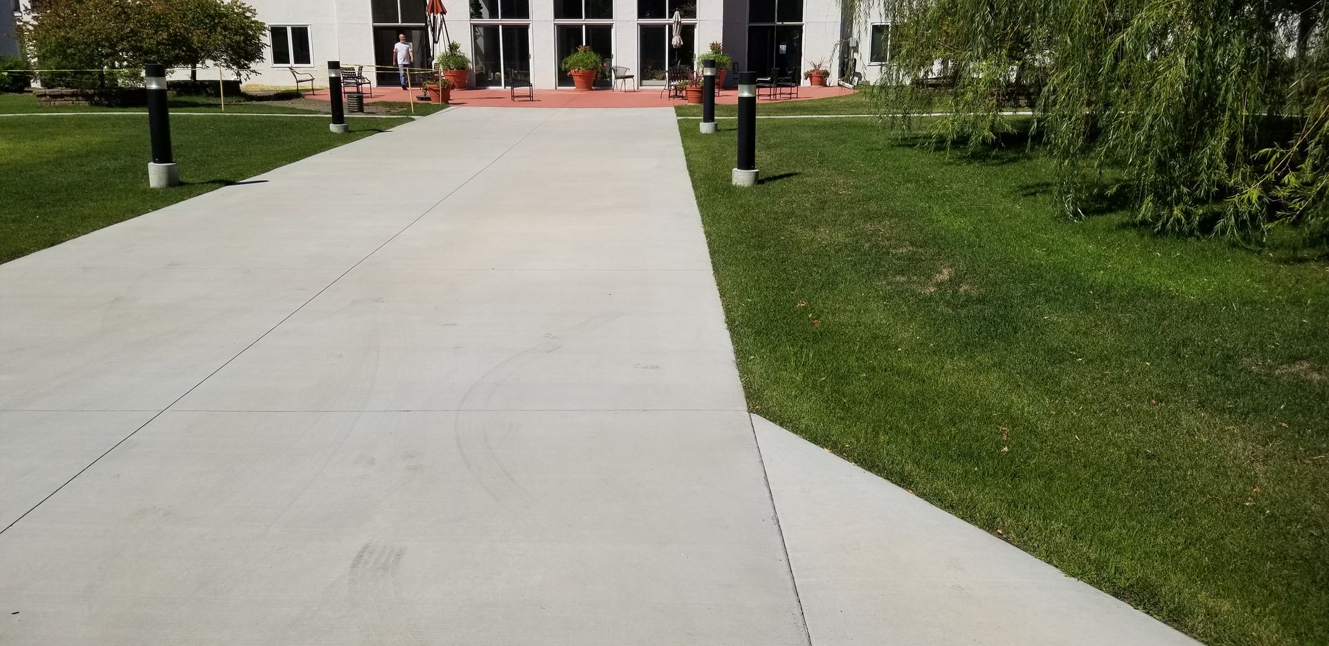 Concrete walkway leading to a building with glass doors, flanked by green grass and bollard lights.