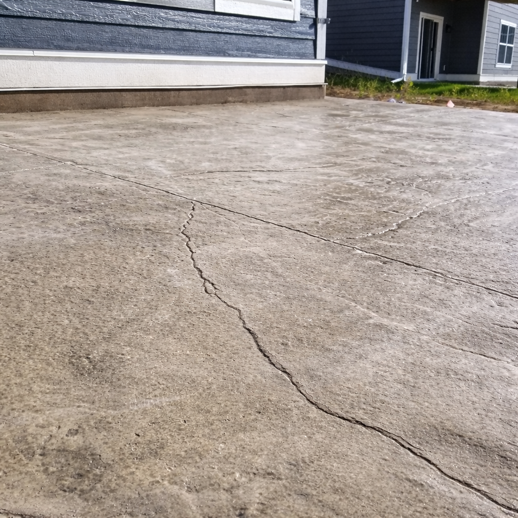 Cracked concrete driveway with a light brown color next to a house with blue siding.