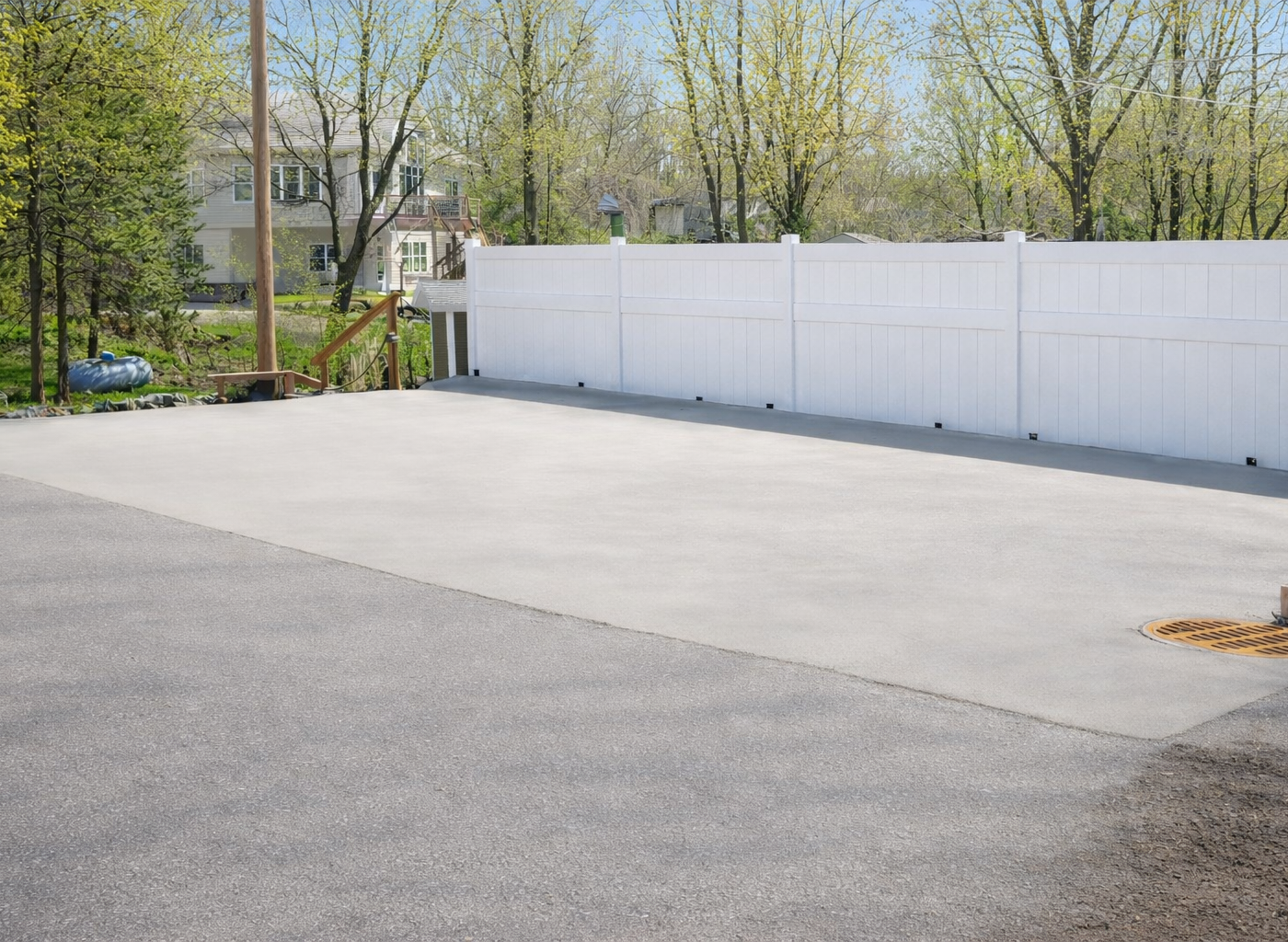 Large paved area with a white fence, trees, and a house in the background.
