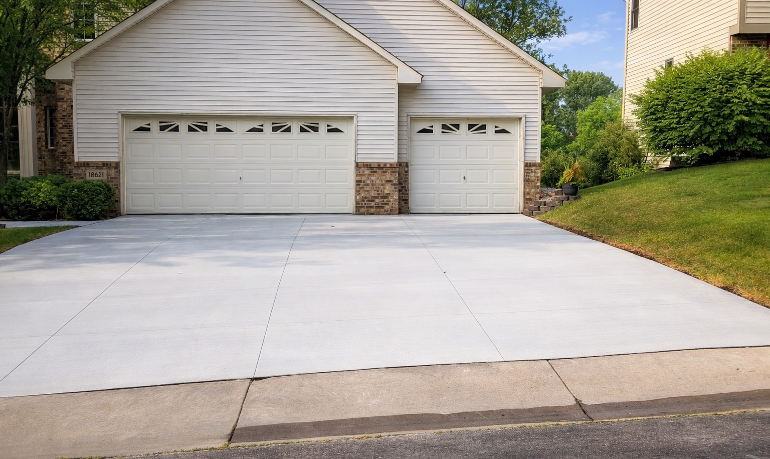 Driveway leading to a two-car garage with white doors, beige stone accents, and a grassy hill to the right.