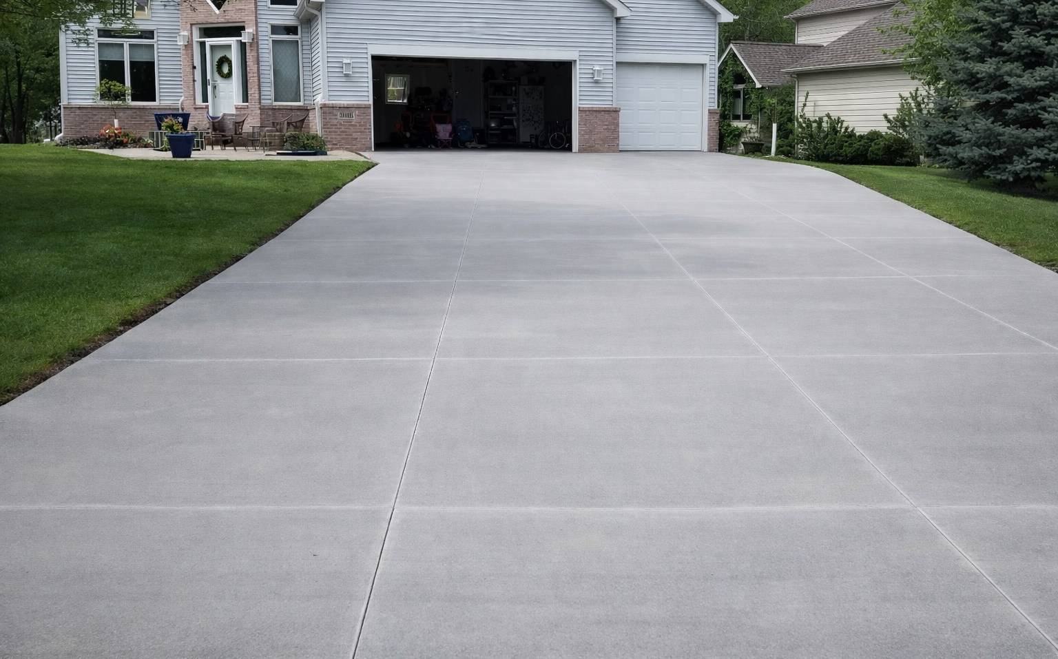 A gray concrete driveway leading to a house with an open garage door and green lawn.