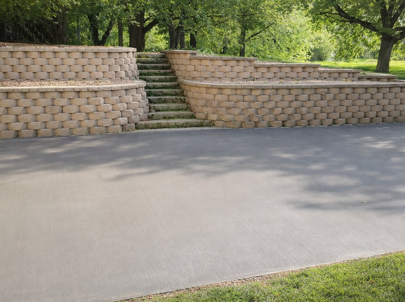 Asphalt driveway leading to terraced beige retaining walls with stairs, grassy area, and trees.