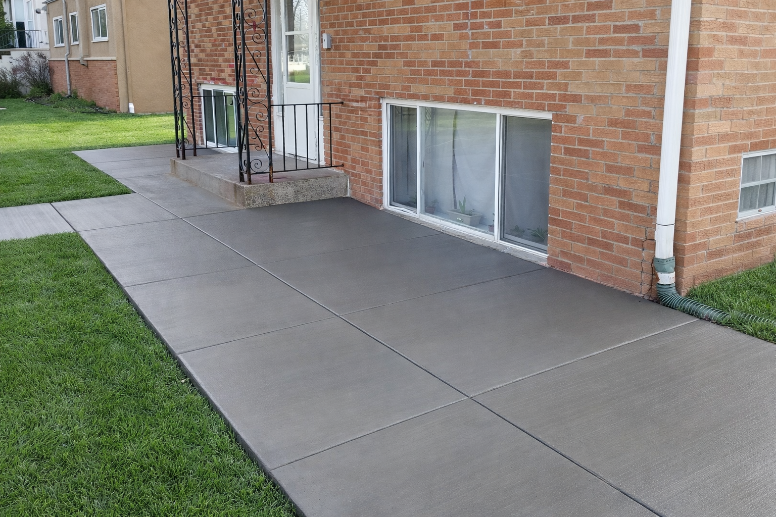 Concrete sidewalk and patio in front of a brick building with windows and green lawn.