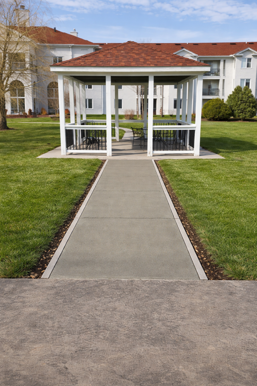 A gazebo with red roof and white pillars sits at the end of a concrete path, surrounded by green grass and buildings.