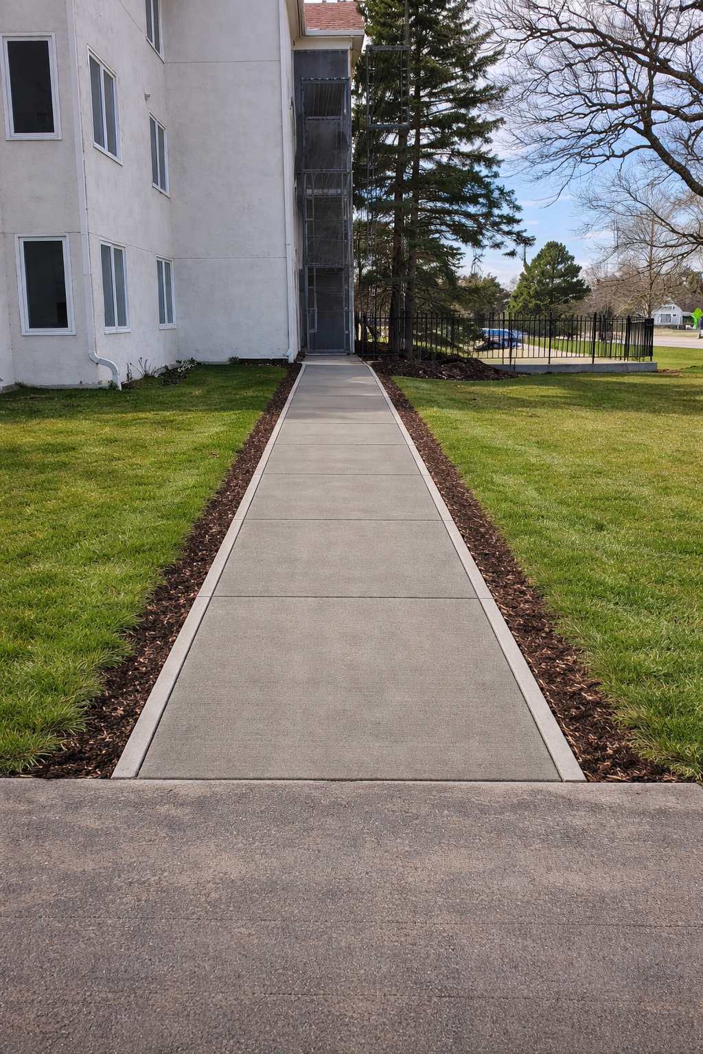 Concrete walkway between a building and a grassy area, with trees and a blue sky in the background.