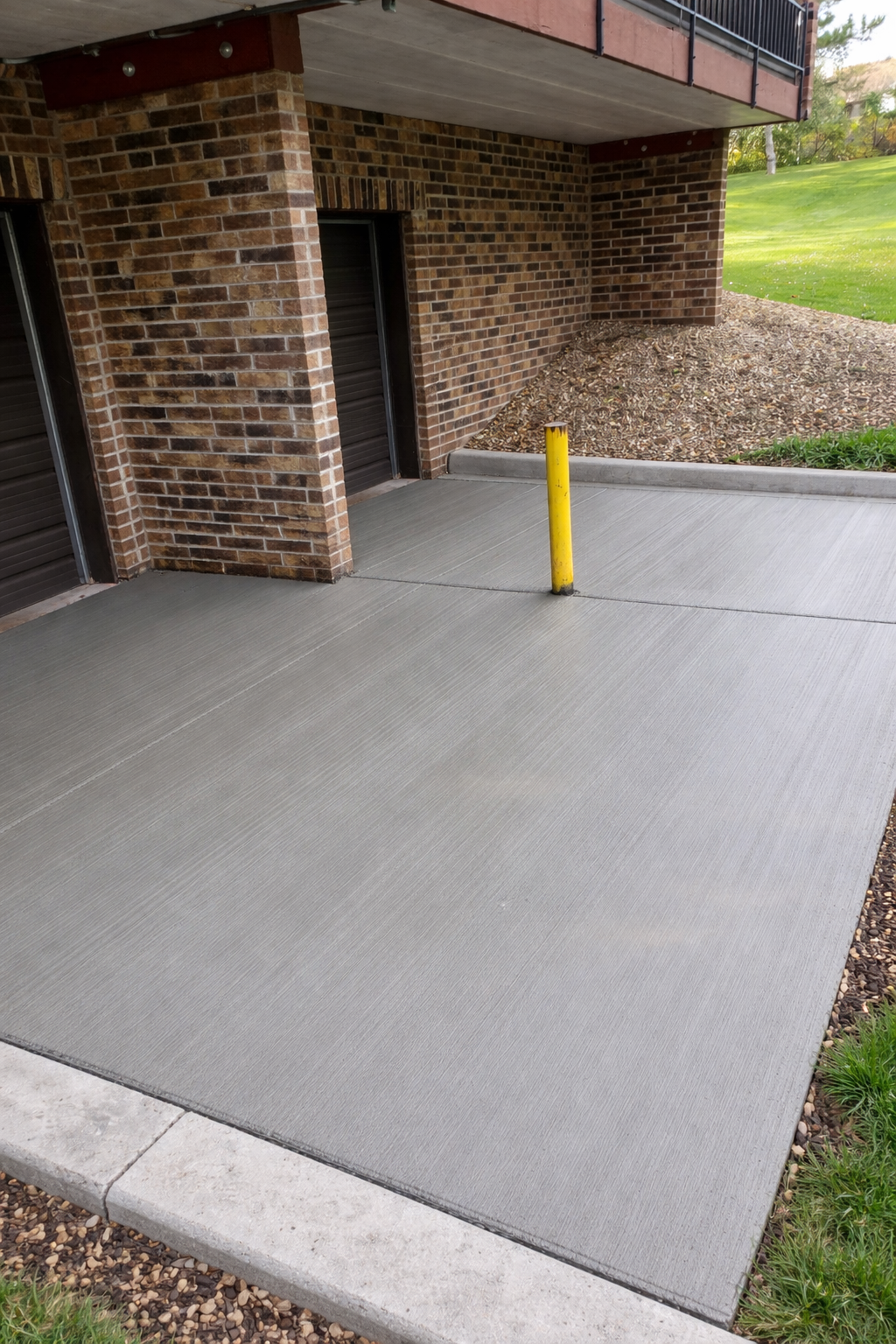 Concrete driveway next to a brick building and a grassy area, with a yellow bollard.