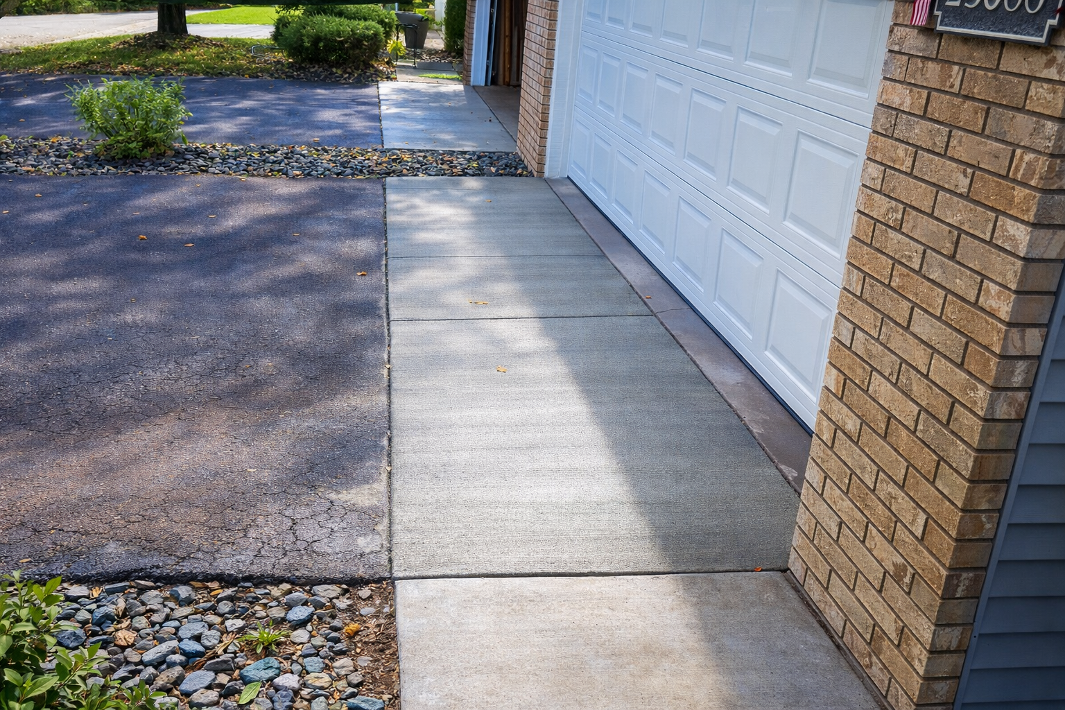 Driveway with asphalt and concrete walkway leading to a garage. Brown brick pillar on right.