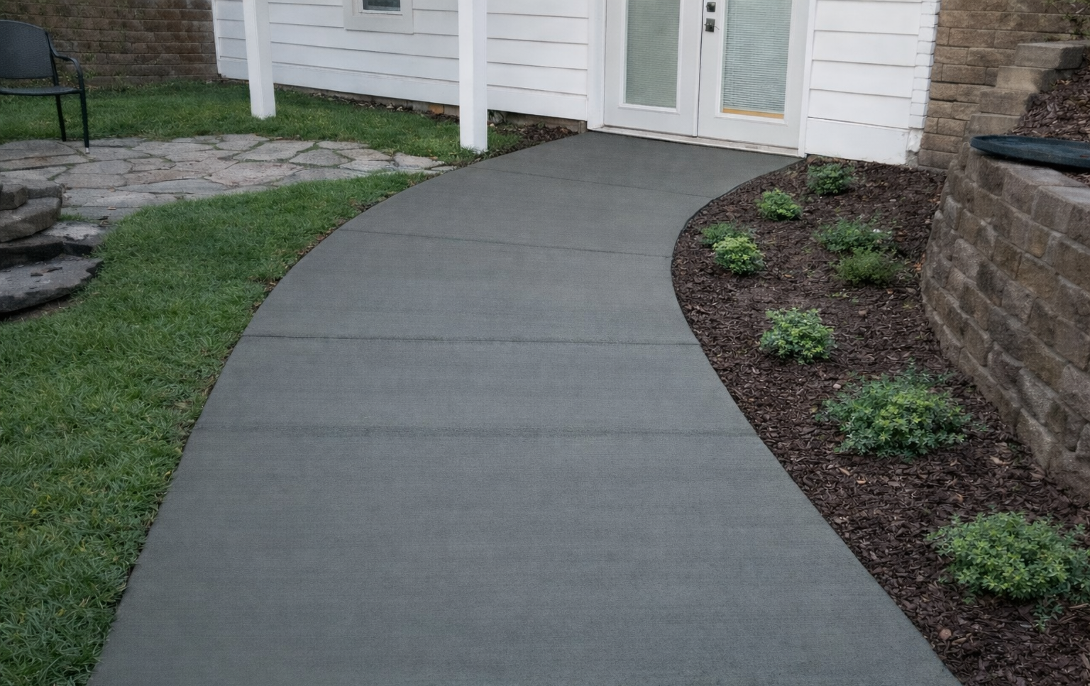 Curving gray concrete walkway leading to white door, flanked by grass and garden beds.