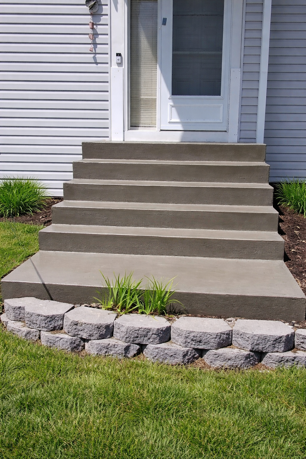 Concrete steps leading up to a white door, framed by light gray siding and a block retaining wall in a grassy yard.