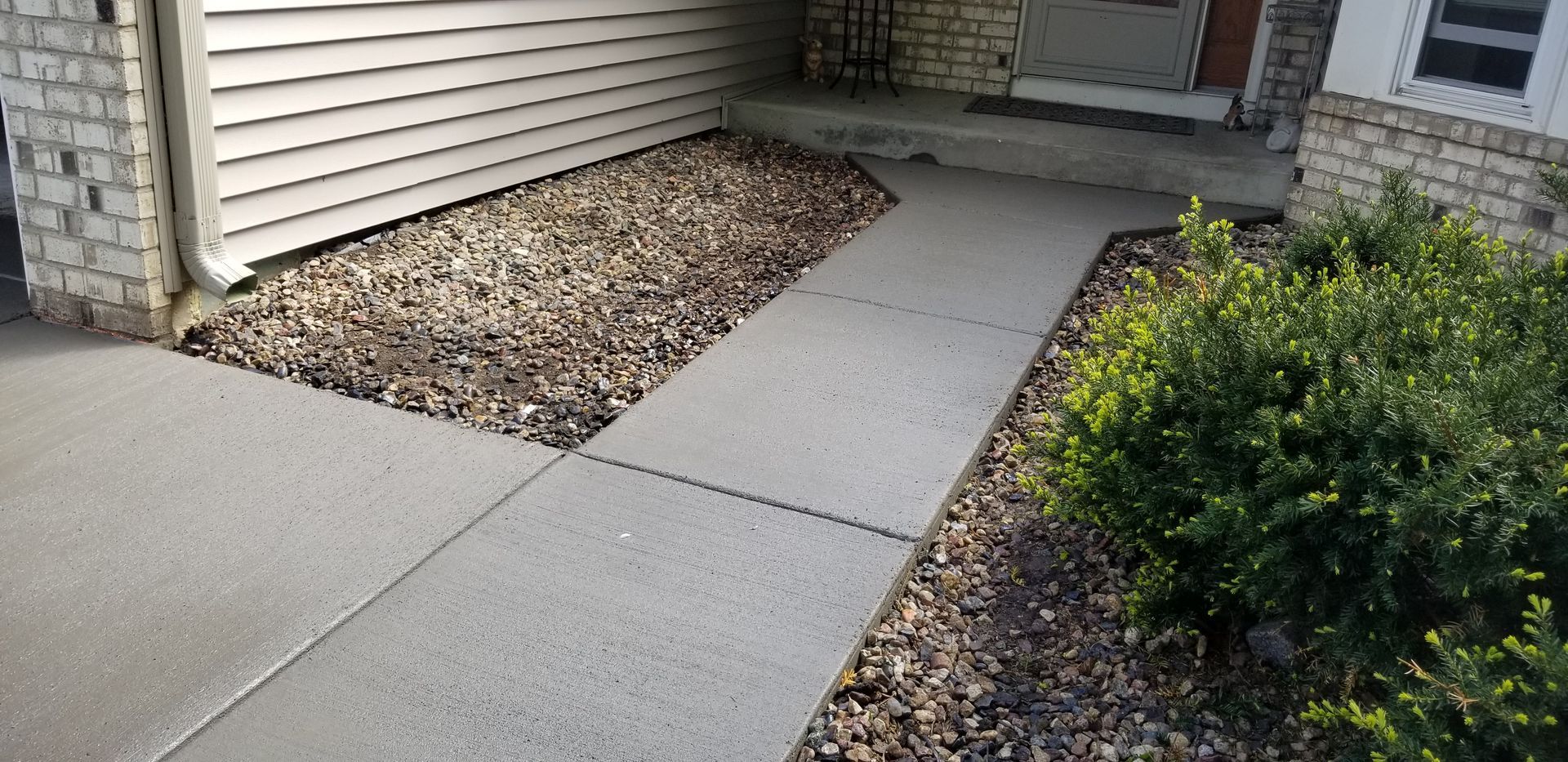 Concrete sidewalk leading to a house entrance, bordered by rocks and a green bush.