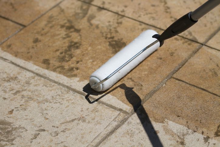 White paint roller applying sealant to tan stone tiles outdoors.