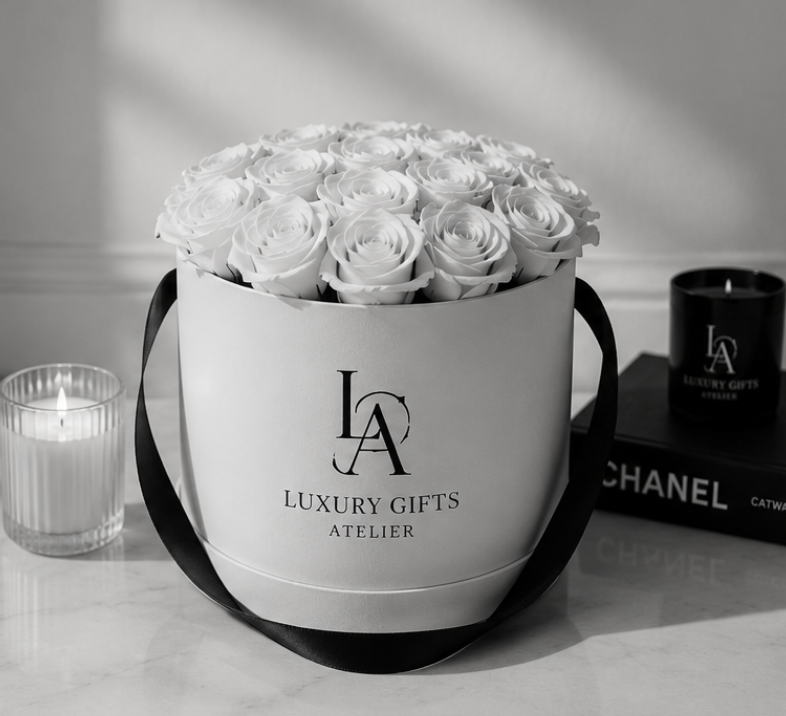 Een boeket witte rozen in een witte geschenkdoos met zwarte handvatten, naast een kaars en Chanel-artikelen op een tafel.
