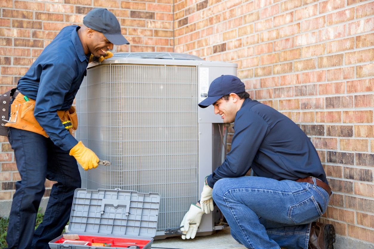 Two men are working on an air conditioner outside of a brick building.
