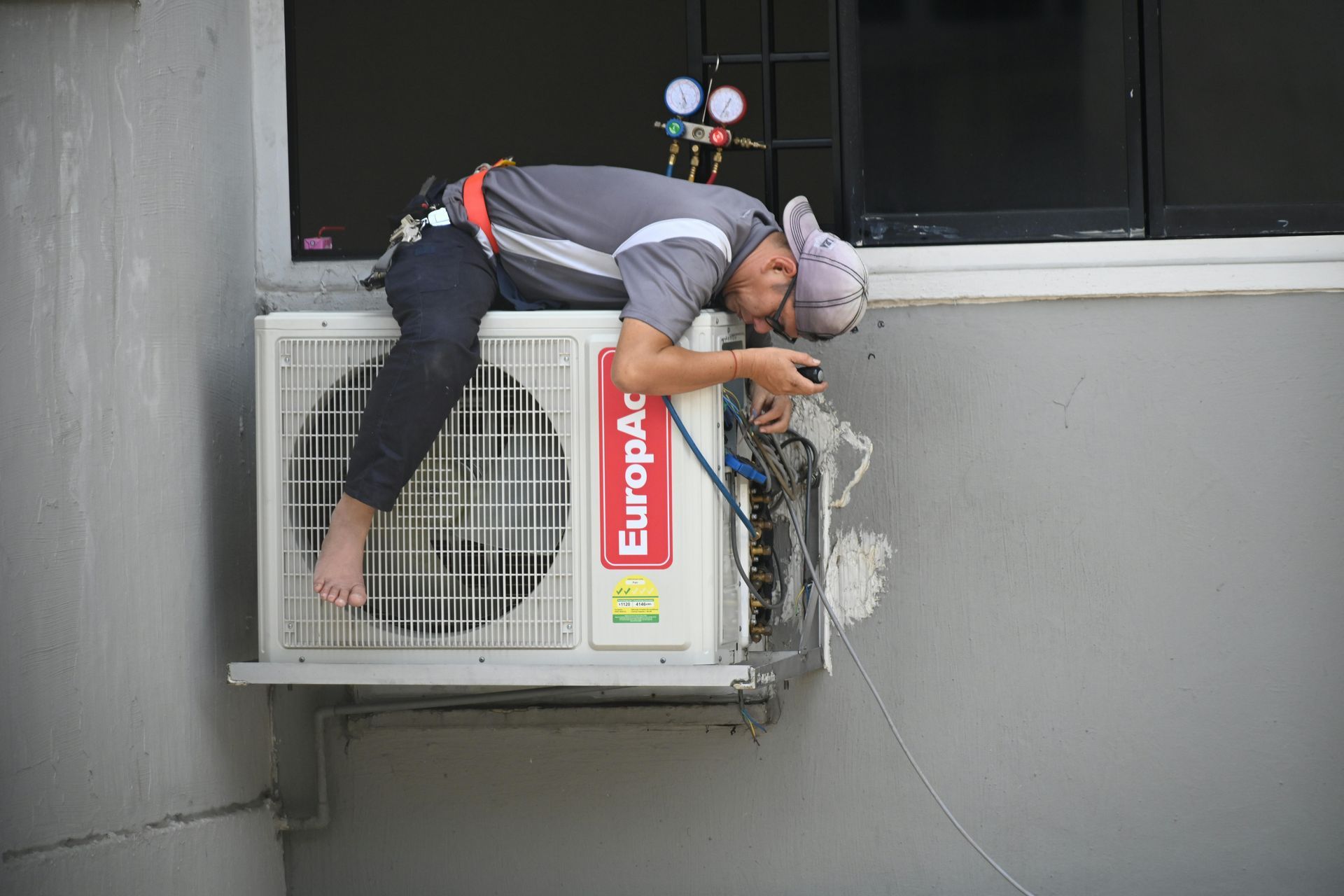 A technician wearing a harness repairs an outdoor air conditioning unit mounted on a grey wall.