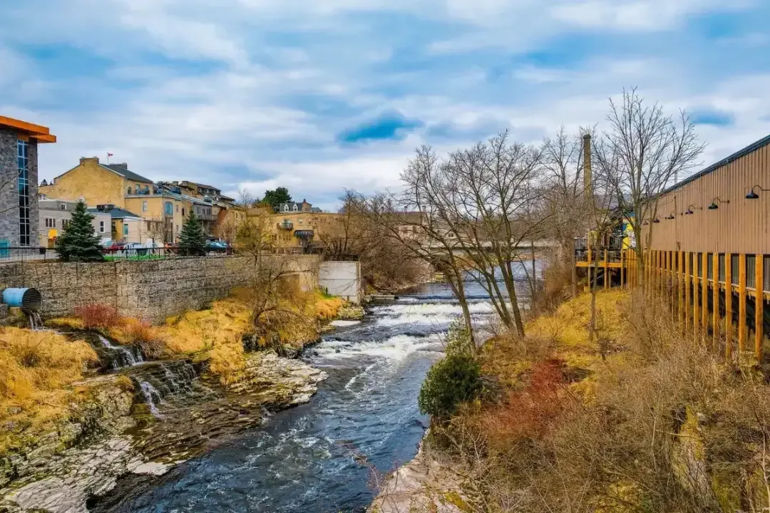 Rushing river beside autumn trees, buildings, and a tan structure under a cloudy sky