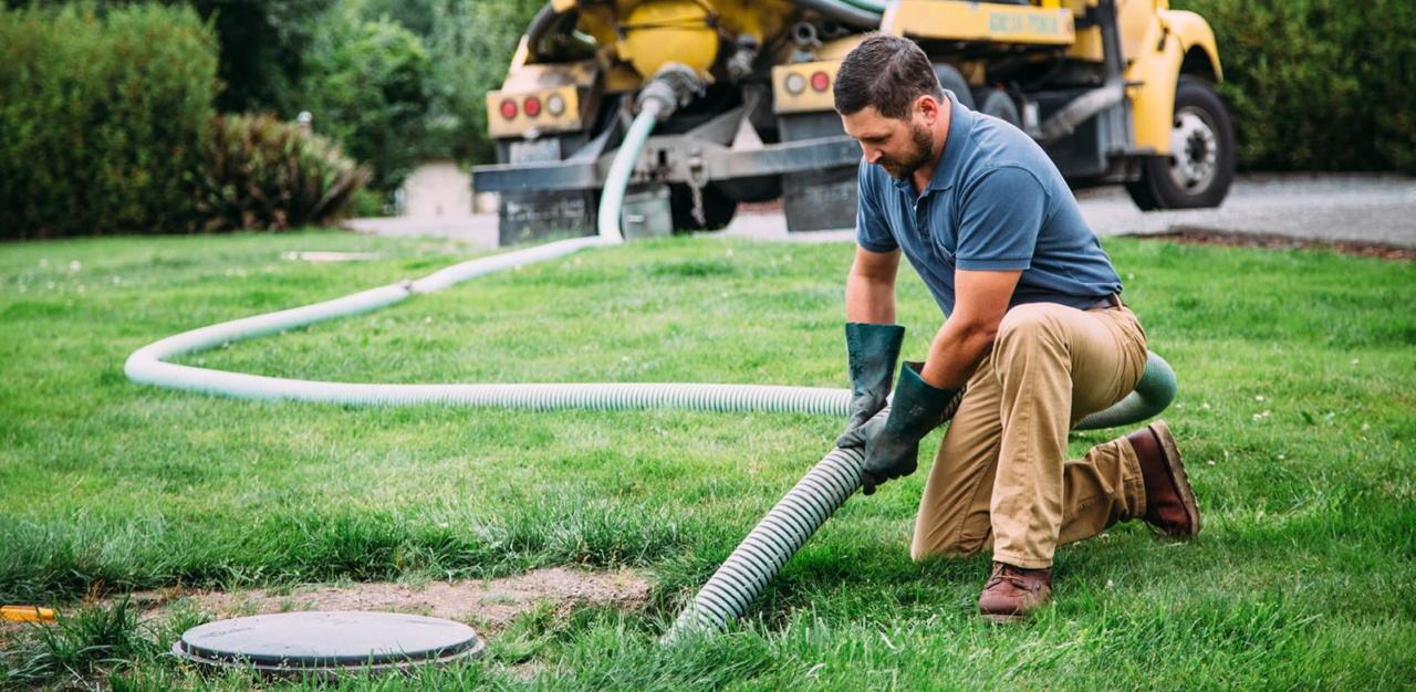 A technician in work clothes kneels on a lawn, guiding a large vacuum hose into an open septic tank lid near a pump truck.
