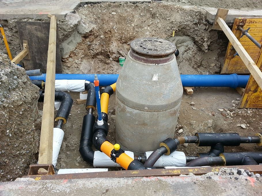 A construction trench showing a concrete manhole surrounded by black-insulated pipes and a blue pipe in the background.