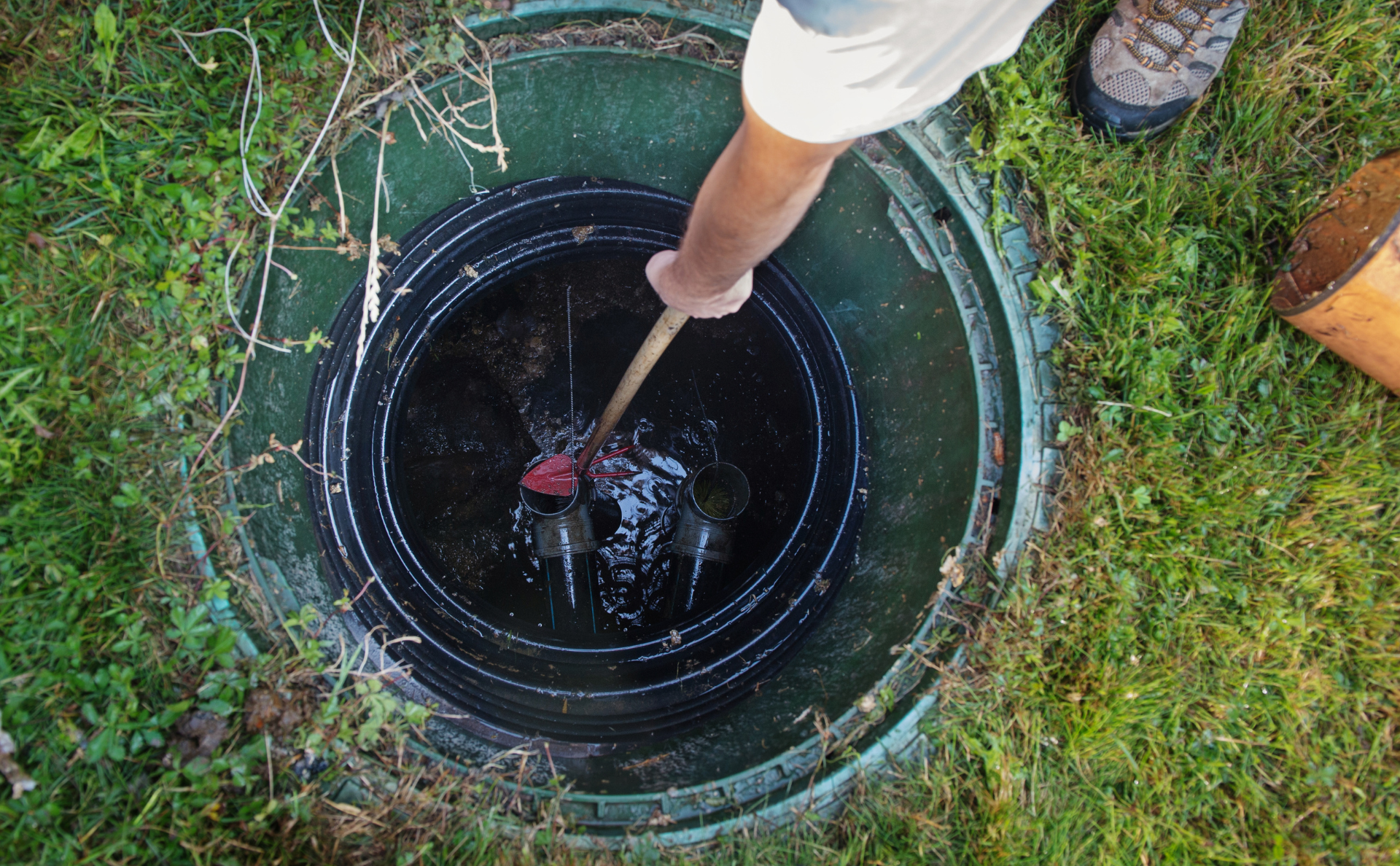 A person uses a pole to inspect or test the liquid levels inside an open septic tank riser in a grassy area.