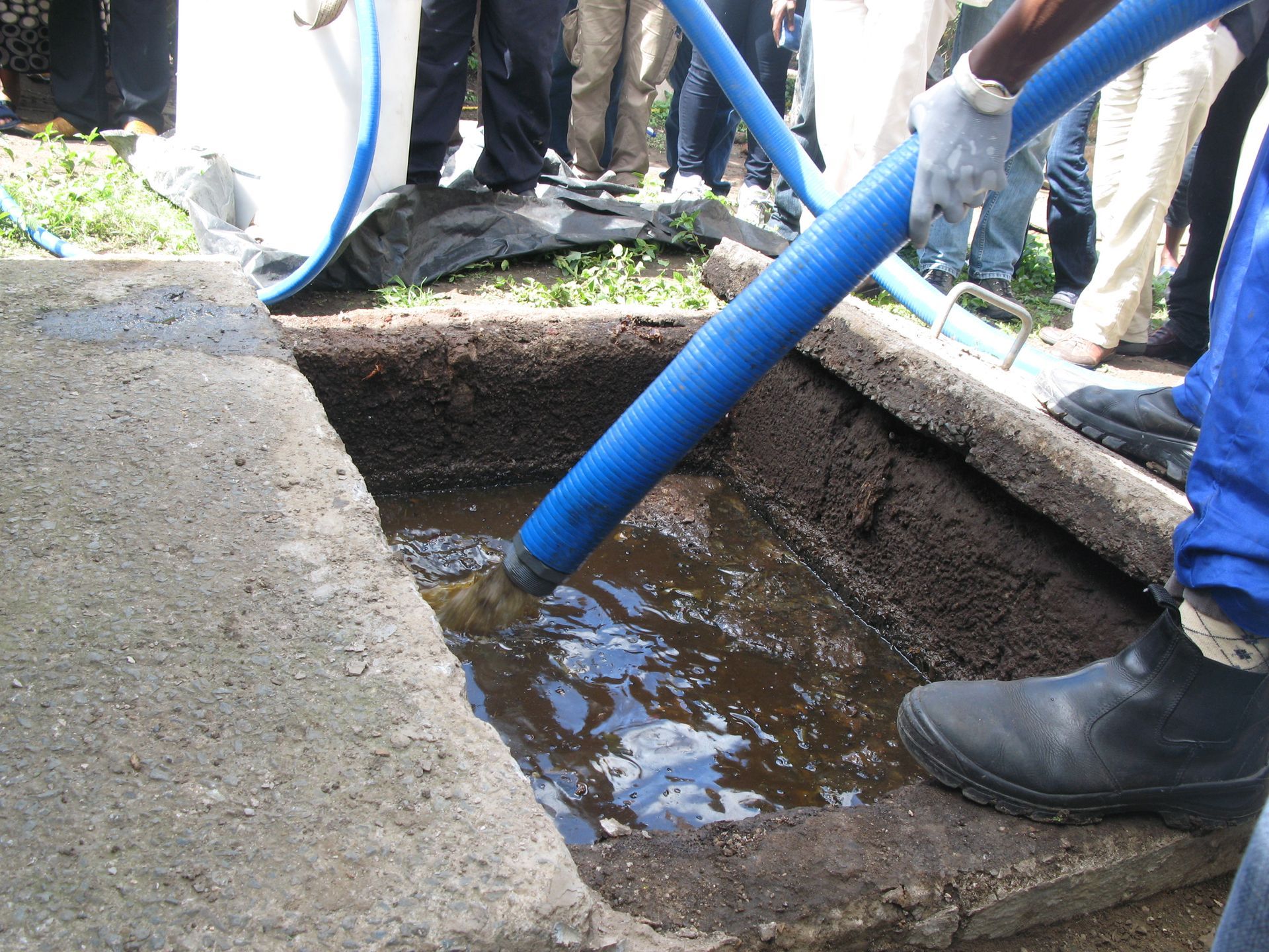 A worker in protective boots and gloves uses a blue hose to pump liquid waste from an open concrete septic tank.