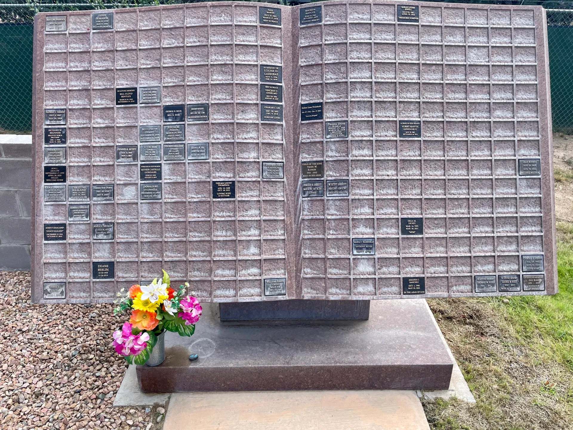 Memorial wall with many small plaques, flowers in front.