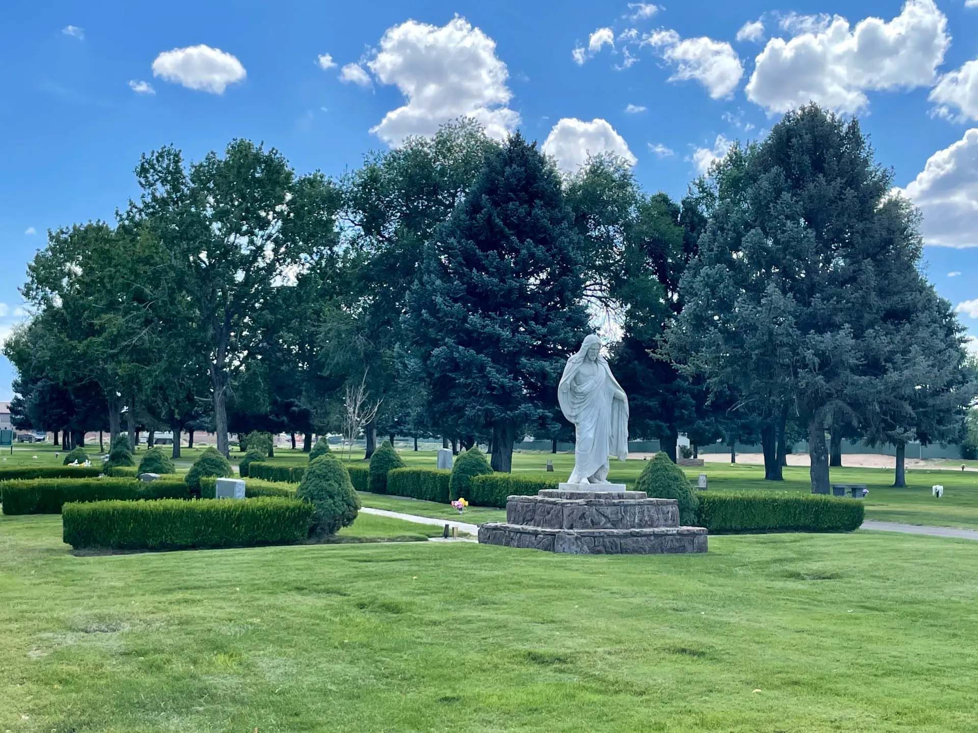Statue in a cemetery, with trees, blue sky, and green grass.