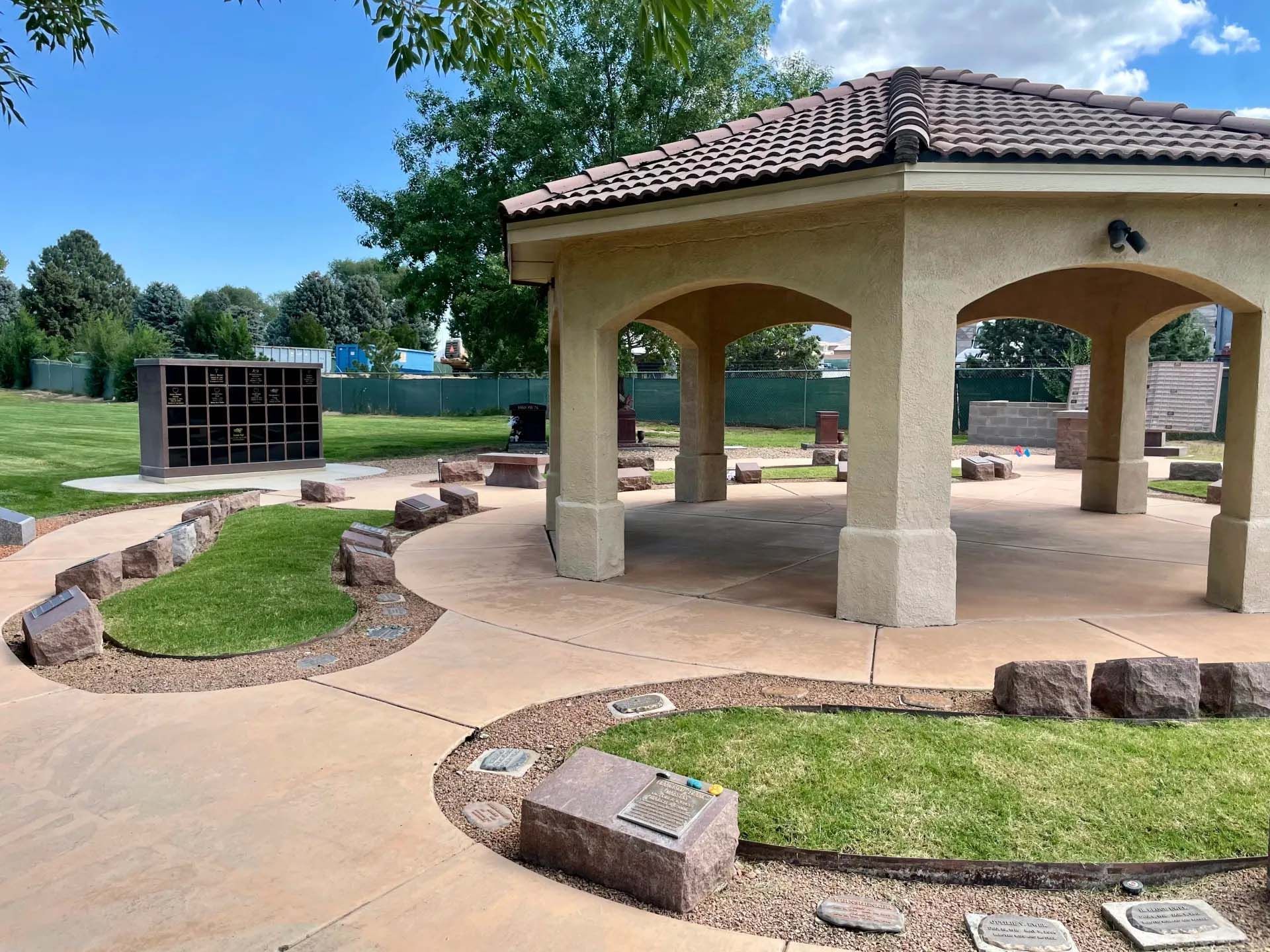 Gazebo with tile roof in park. Pathways with rocks and greenery lead to memorial structures. Sunny day.