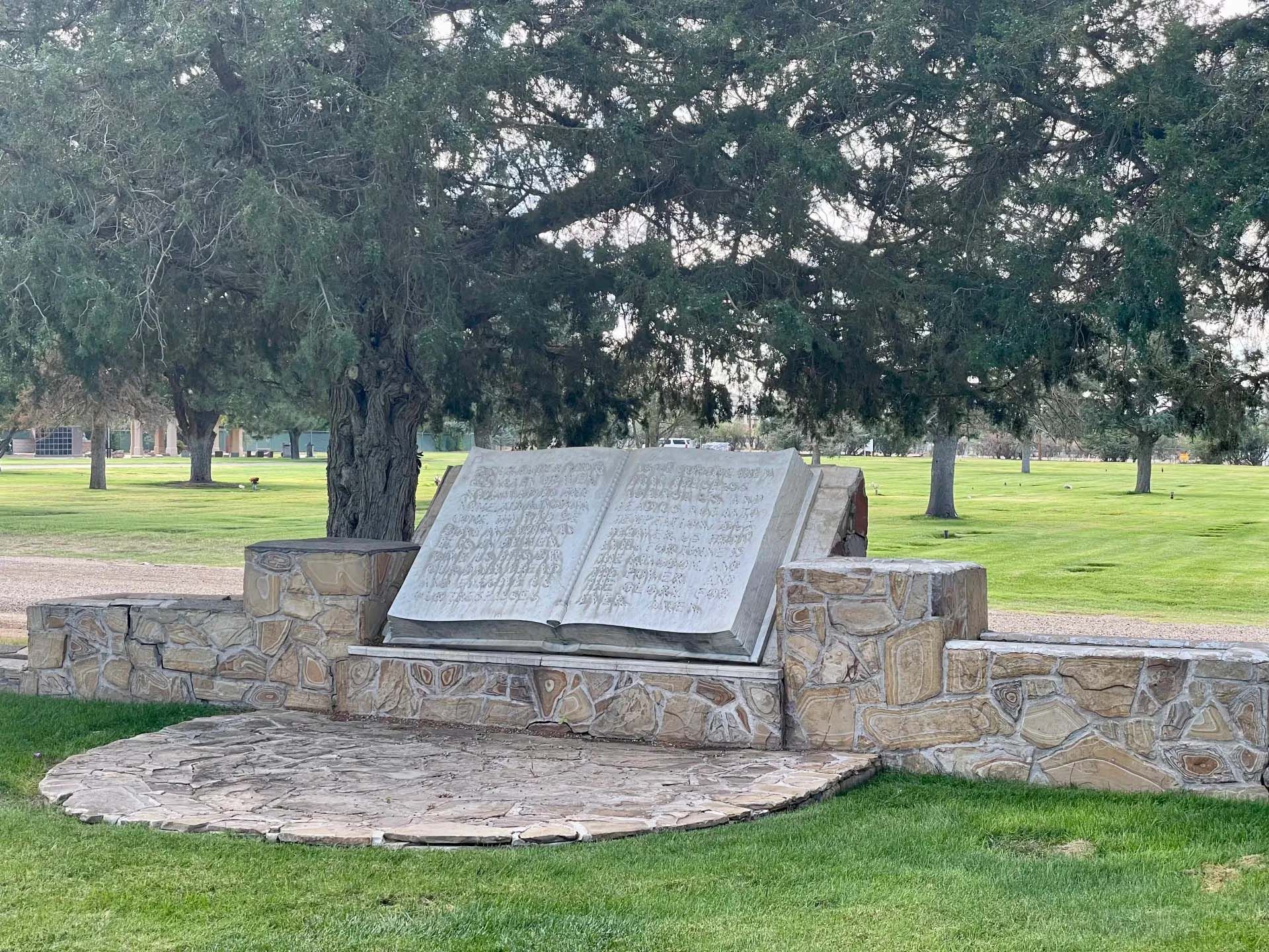 Stone book-shaped memorial on a stone base, set in a grassy park, with trees in the background.