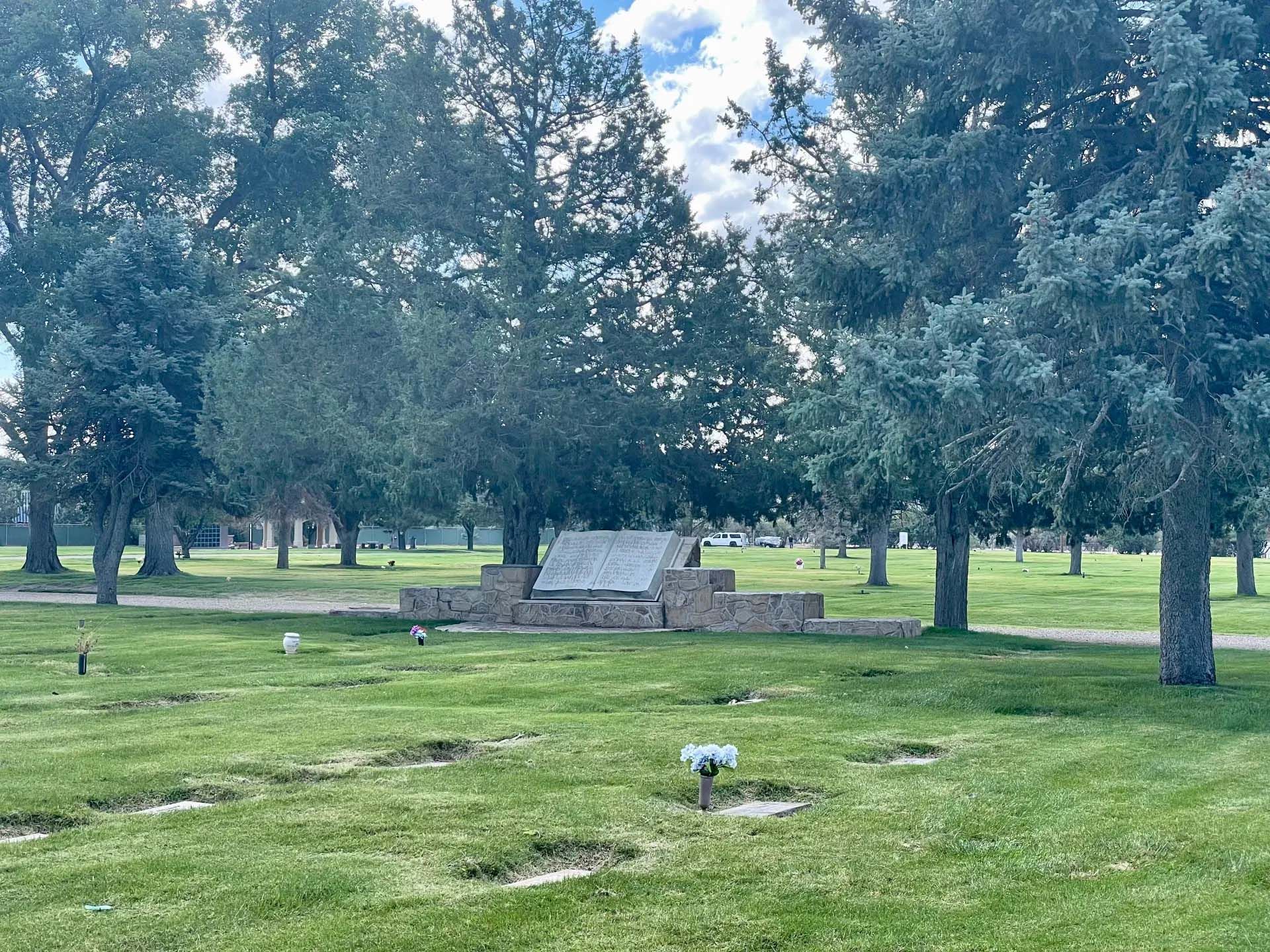Grave memorial in a grassy cemetery surrounded by trees under a cloudy sky.