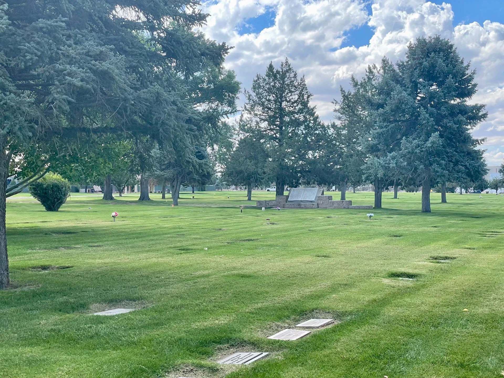 Grassy cemetery under a partly cloudy sky, with trees and headstones scattered throughout.